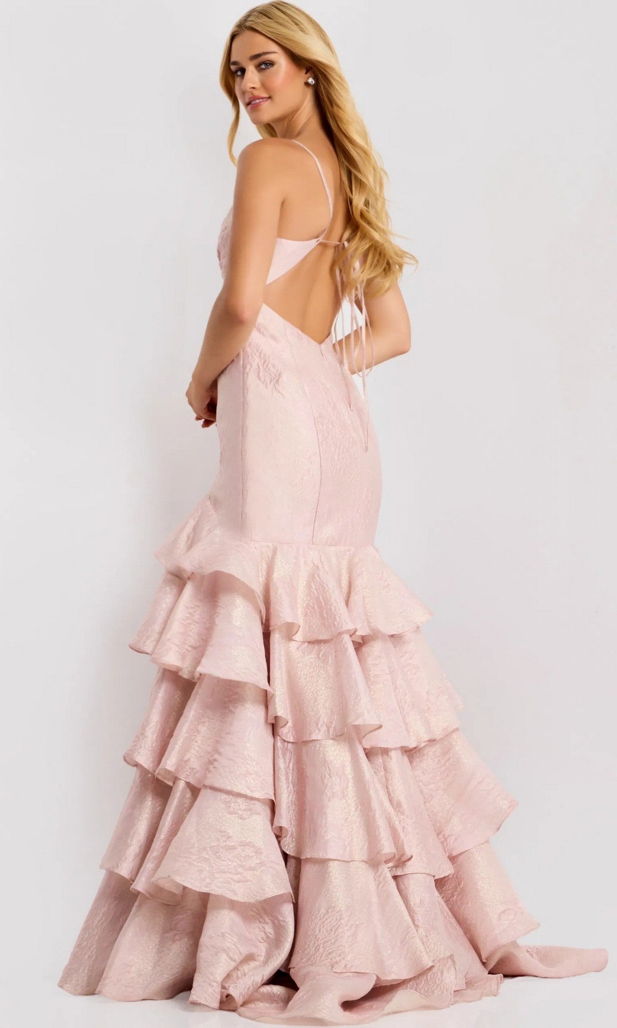 A woman models the Jovani 47329 long prom dress in pale pink, featuring an open back and tiered ruffle skirt, posing slightly away to showcase the gown’s elegant details against a plain background.